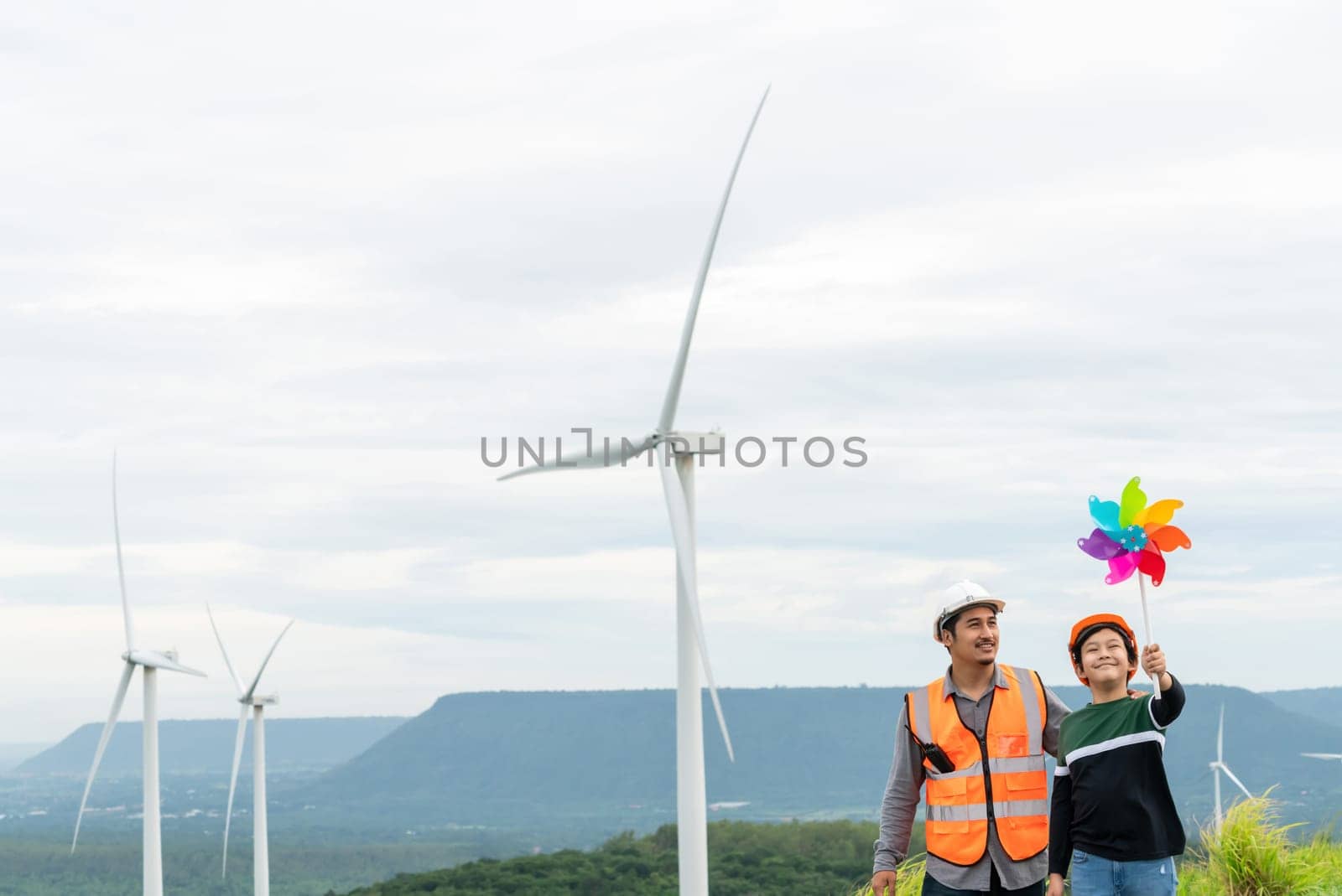 Engineer with his son holding windmill toy on a wind farm atop a hill or mountain. Progressive ideal for the future production of renewable, sustainable energy. Energy generated from wind turbine.