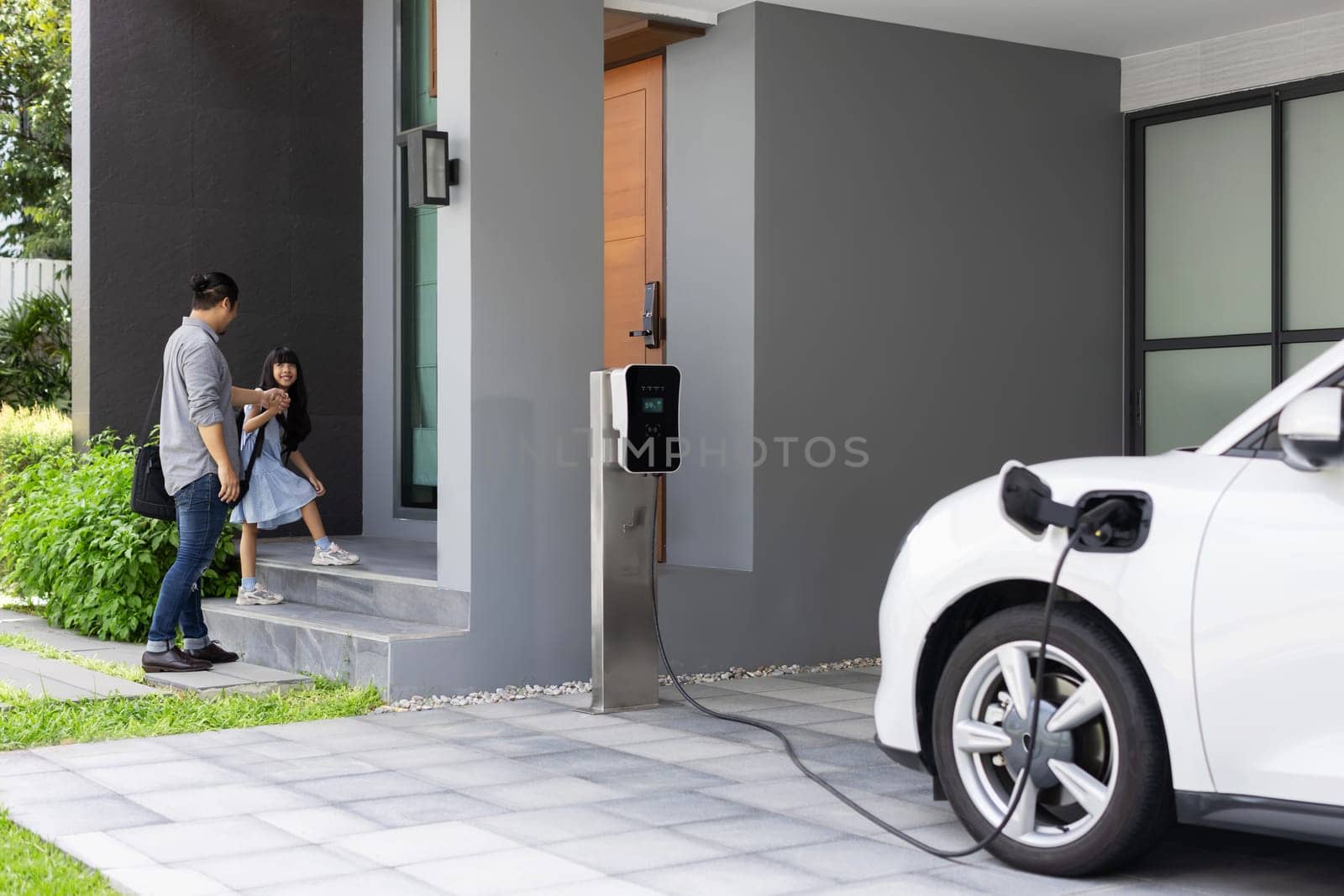 Progressive father and daughter plugs EV charger from home charging station to electric vehicle. Future eco-friendly car with EV cars powered by renewable source of clean energy.