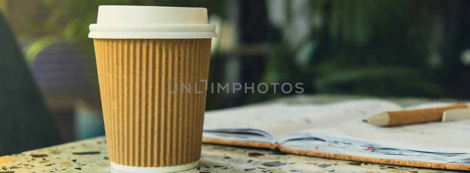 Hot latte coffee in craft recycling paper cup with paper notebook. A take away paper cup on cafe table. Freelance Workspace notebook with coffee for productive work and study. Working comfortably. Empty workspace. Blurred background. Mockup Coffee break. Online job or studying co-working space