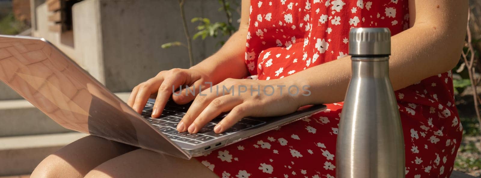 Unrecognizable Young woman in red dress drinking water from reusable metal bottle with laptop. Freelancer's place of work. Study and work online. Remote business education. View webinar. E-learning Workstation on wooden bench. Mockup Coffee break. Making plans for next week year. Being mindful reducing stress and slowing-down