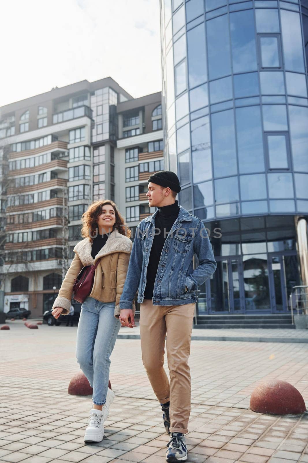 Cheerful couple in casual warm clothes have a walk outdoors in the city near business building.