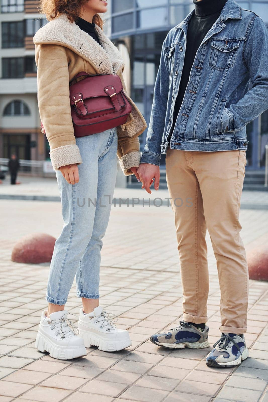 Cheerful couple in casual warm clothes have a walk outdoors in the city near business building by Standret