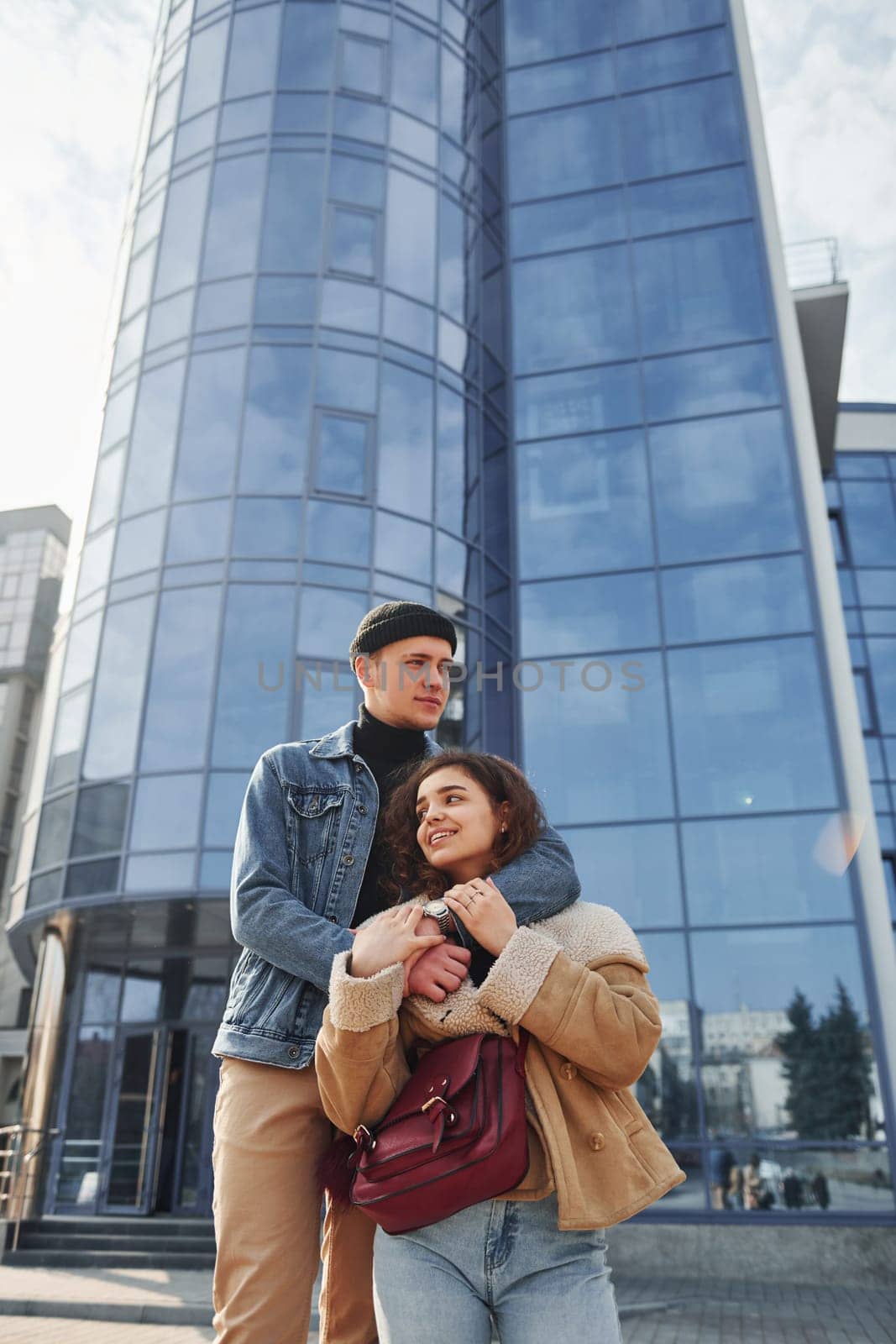 Cheerful couple in casual warm clothes have a walk outdoors in the city near business building by Standret