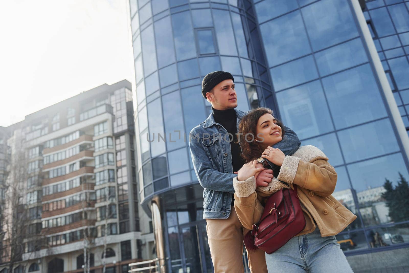 Cheerful couple in casual warm clothes have a walk outdoors in the city near business building by Standret