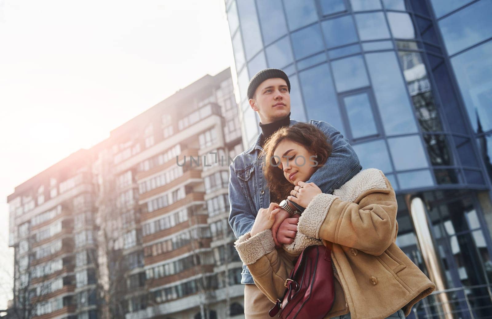 Cheerful couple in casual warm clothes have a walk outdoors in the city near business building by Standret