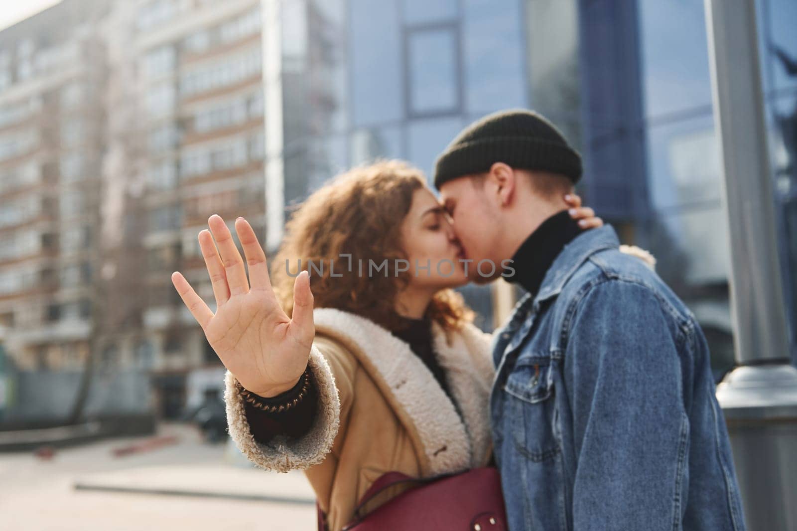 Cheerful couple in casual warm clothes kissing outdoors in the city near business building.