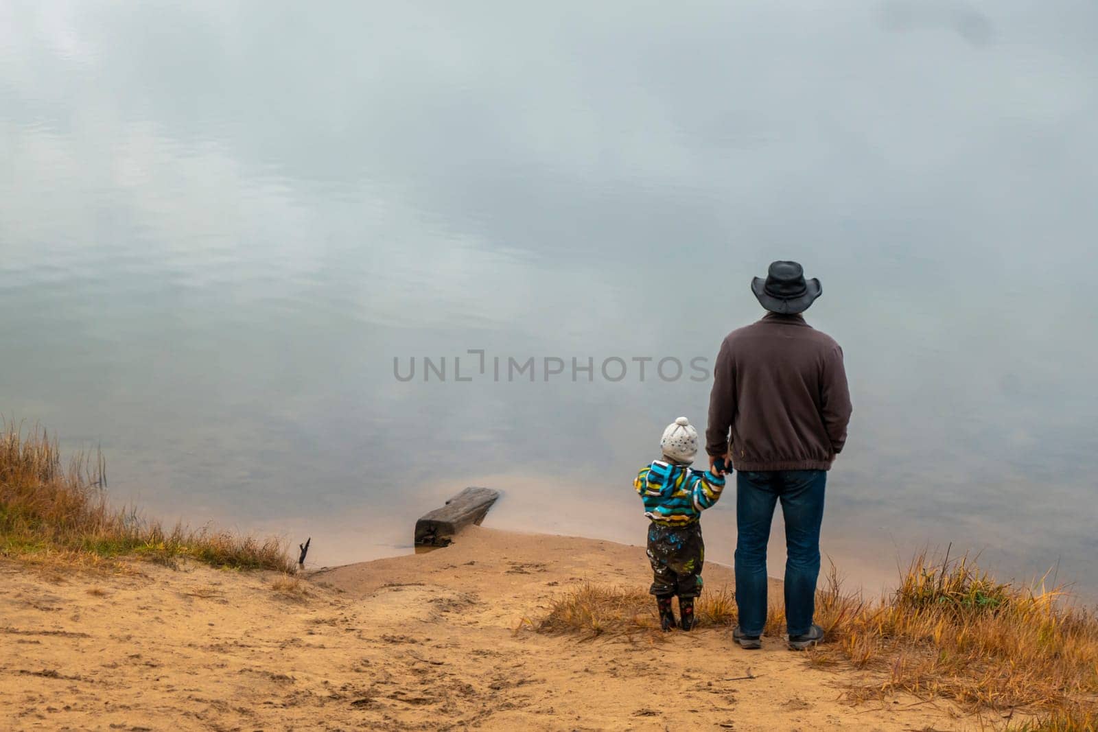 A happy family of two is standing on a cloudy autumn day on the bank of the lake and looking into the distance at the water surface, son and father stand with their backs to the camera
