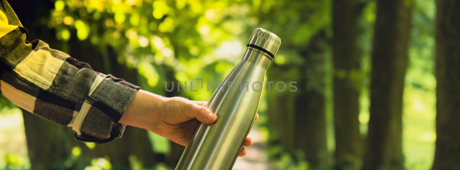 Close-up of Unrecognizable female hand holding Water bottle. Reusable steel thermo water bottle in park. Sustainable lifestyle. Plastic free zero waste free living. Go green Environment protection. Woman drinking water in park Banner size