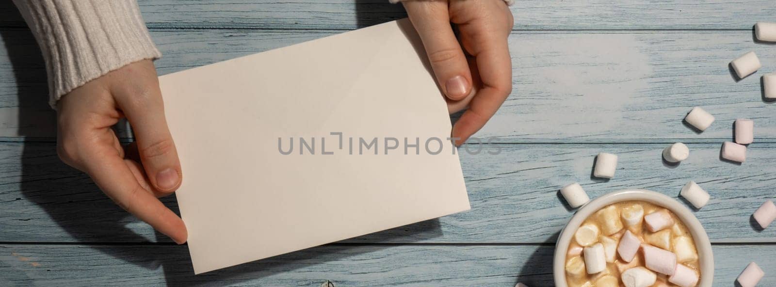 Banner Female hands holding empty greeting or invitation card mock up with white cup of coffee and marshmallows on wooden blue background. Blank paper copy space for your text. Valentines day holiday. Top view, flat lay minimalist branding concept