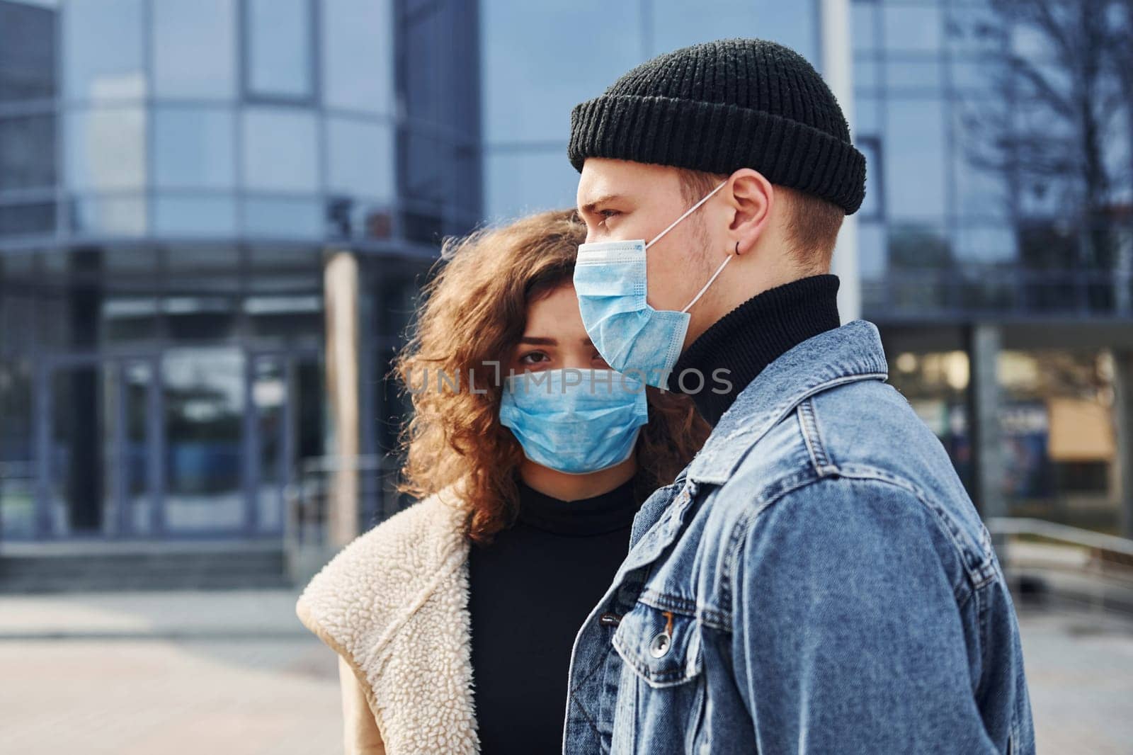 Couple in protective masks have a walk outdoors in the city near business building at quarantine time. Conception of coronavirus.