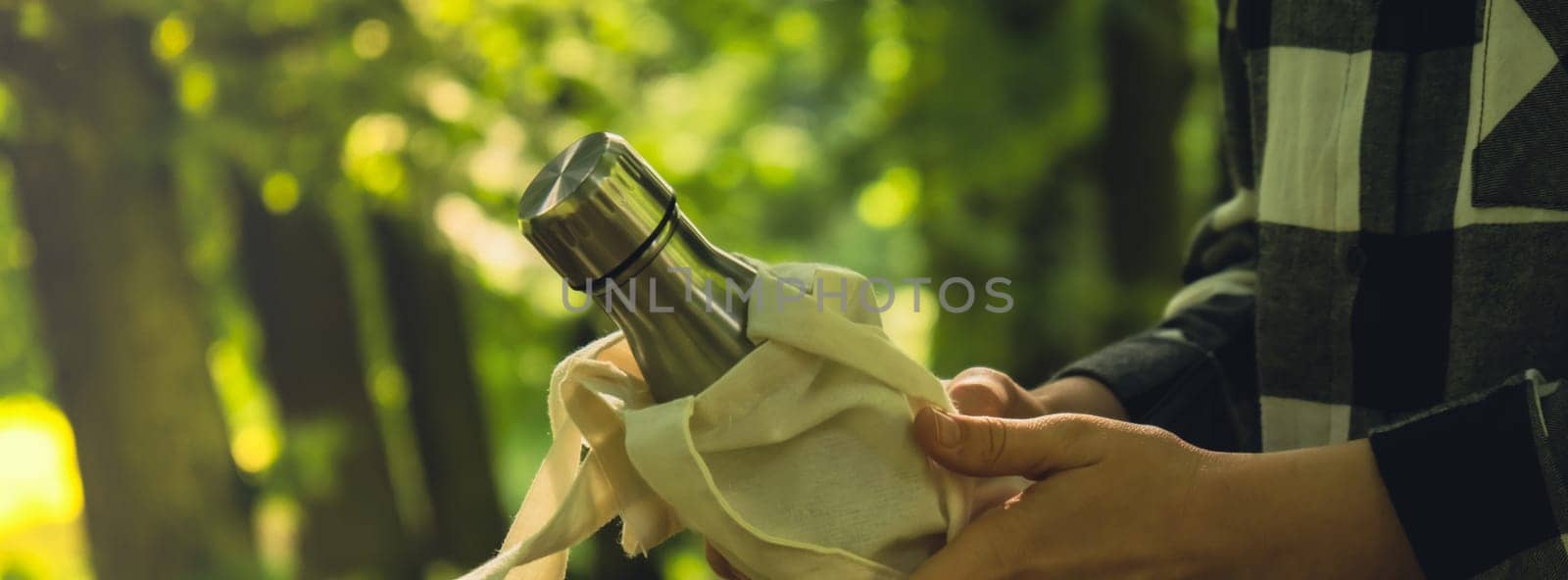 Close-up of Unrecognizable female hand holding Water bottle from eco bag. Reusable steel thermo water bottle in park. Sustainable lifestyle. Plastic free zero waste free living. Go green Environment protection. Woman drinking water in park Banner size