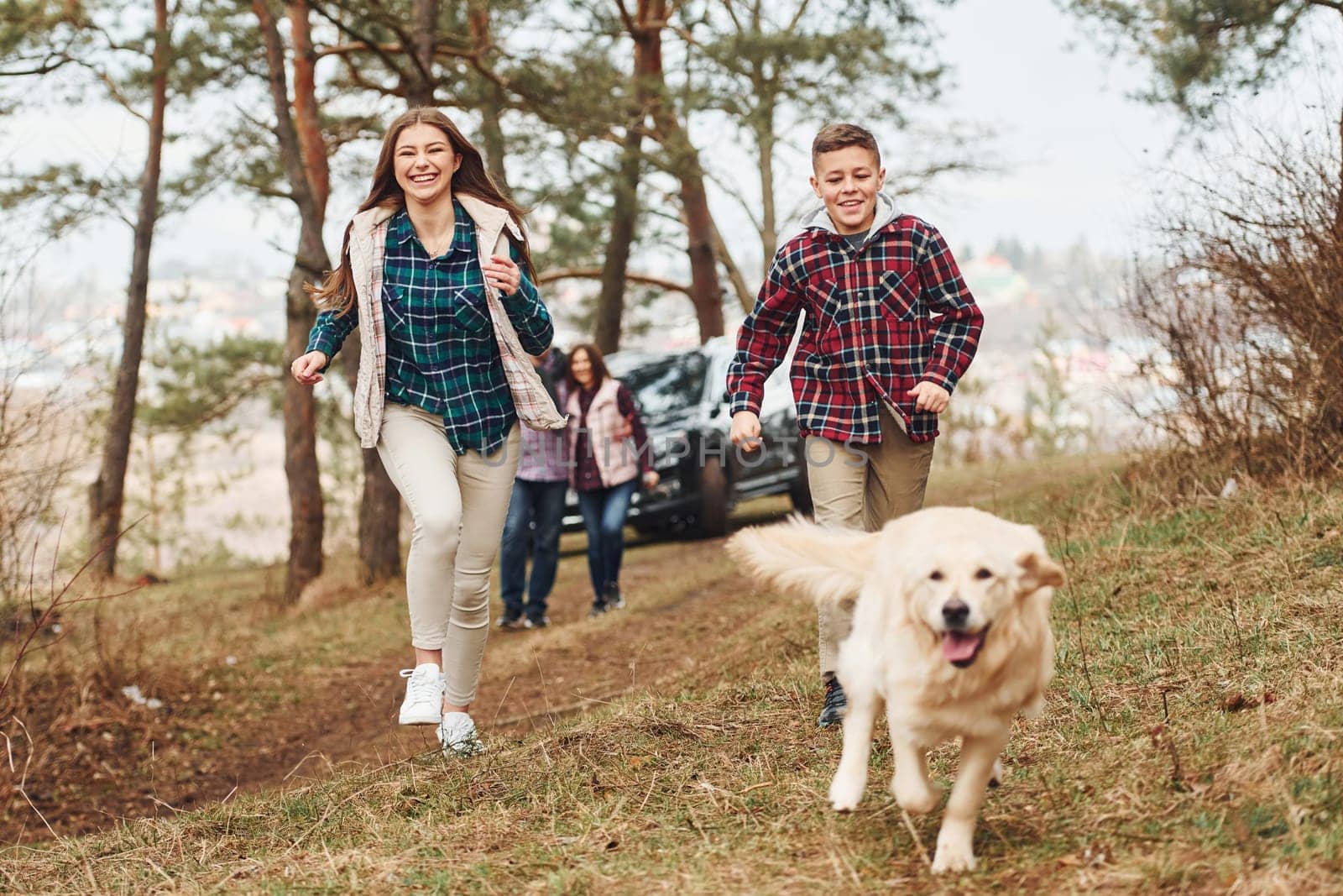 Happy family runs and having fun with their dog near modern car outdoors in forest.