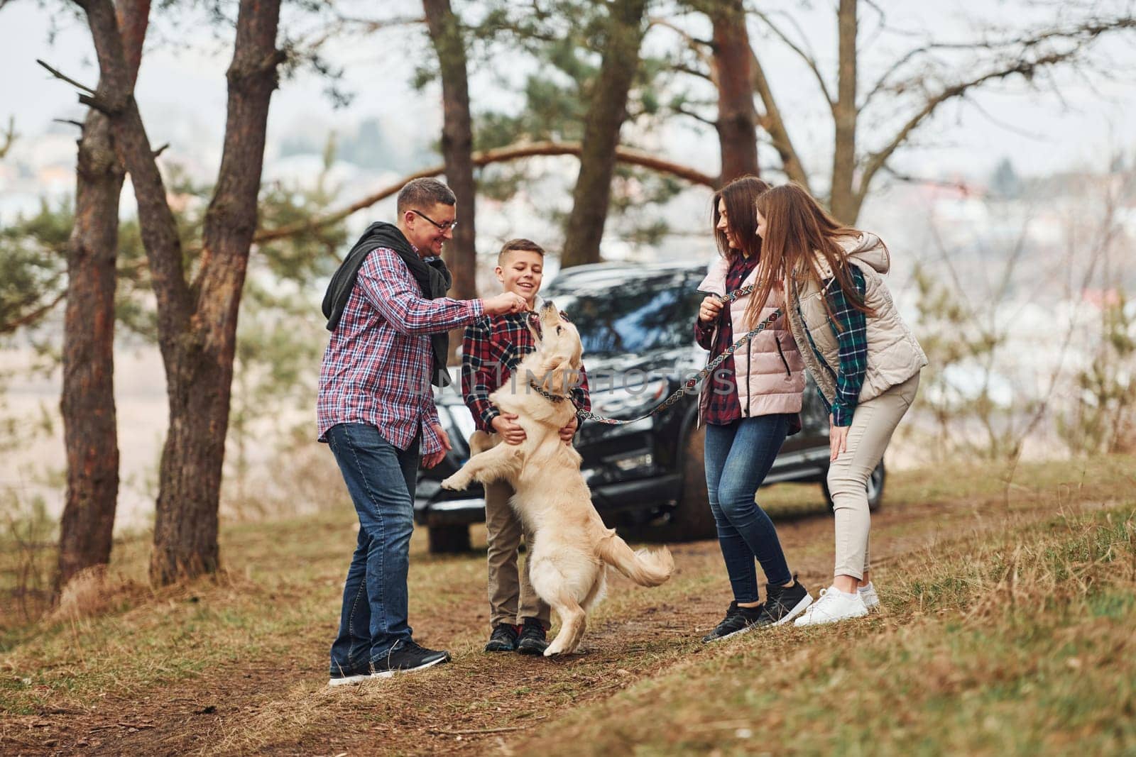 Happy family have fun with their active dog near modern car outdoors in forest.