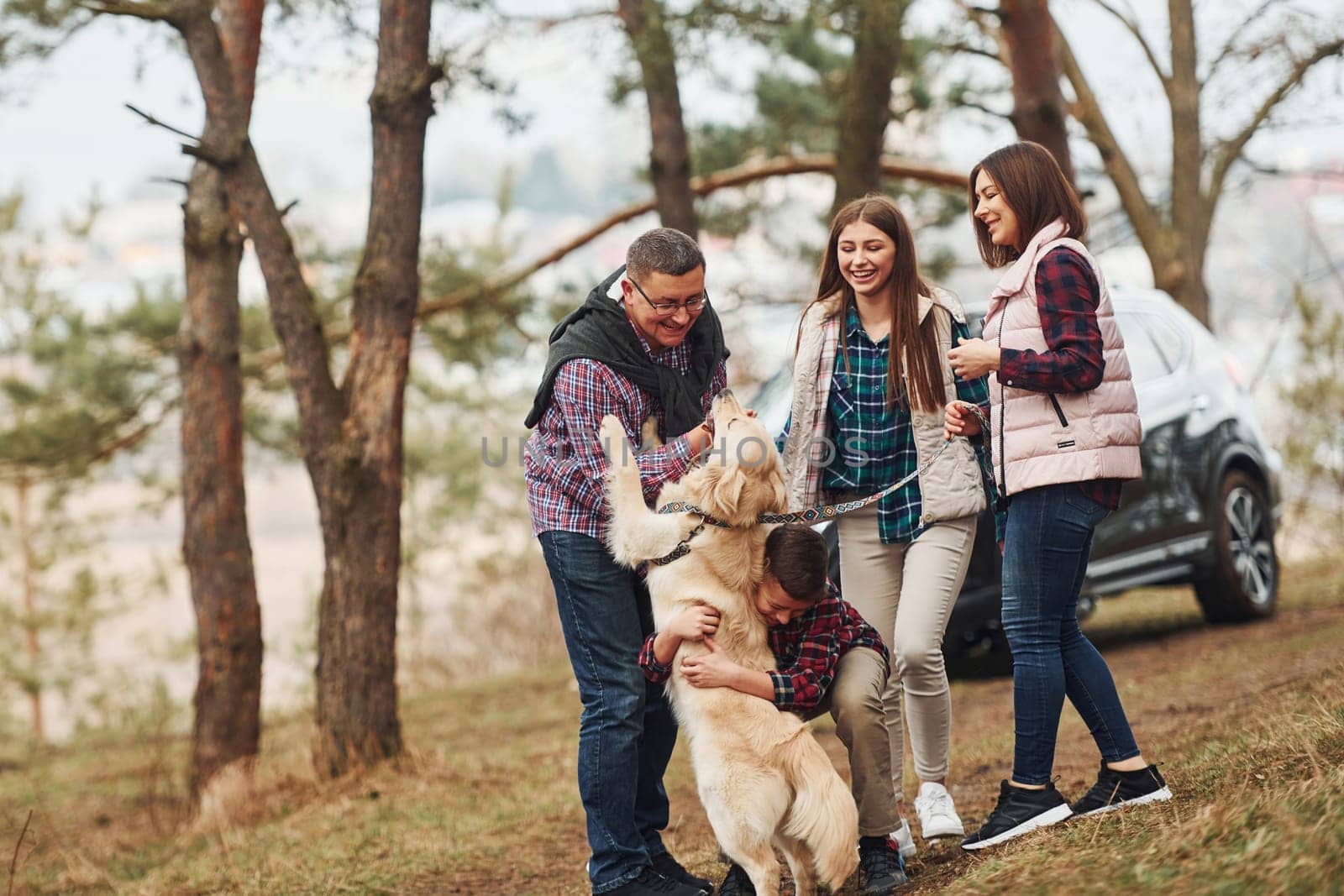 Happy family have fun with their active dog near modern car outdoors in forest.