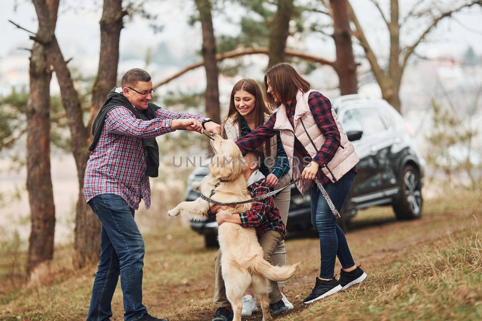 Happy family have fun with their active dog near modern car outdoors in forest.