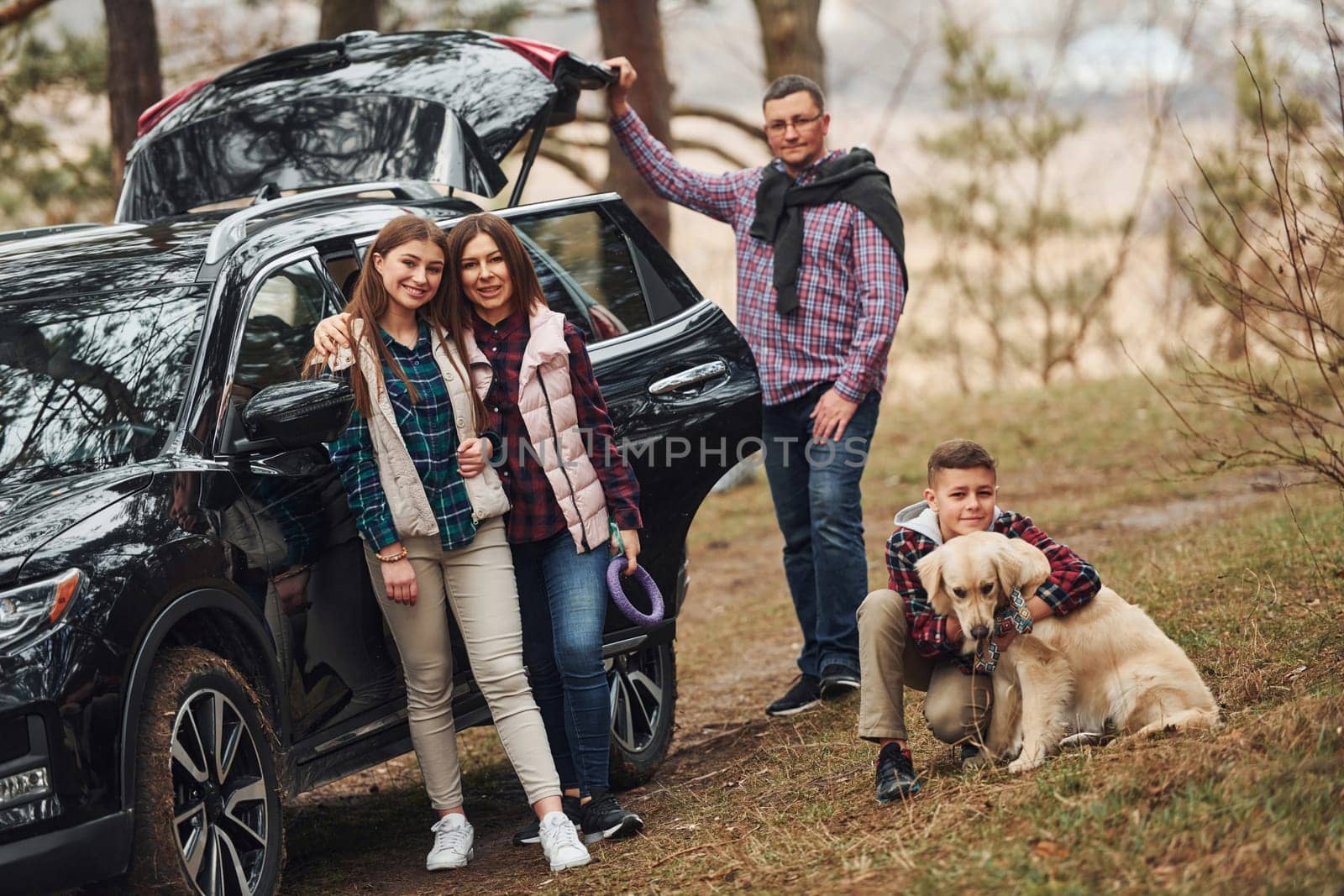 Happy family have fun with their dog near modern car outdoors in forest.