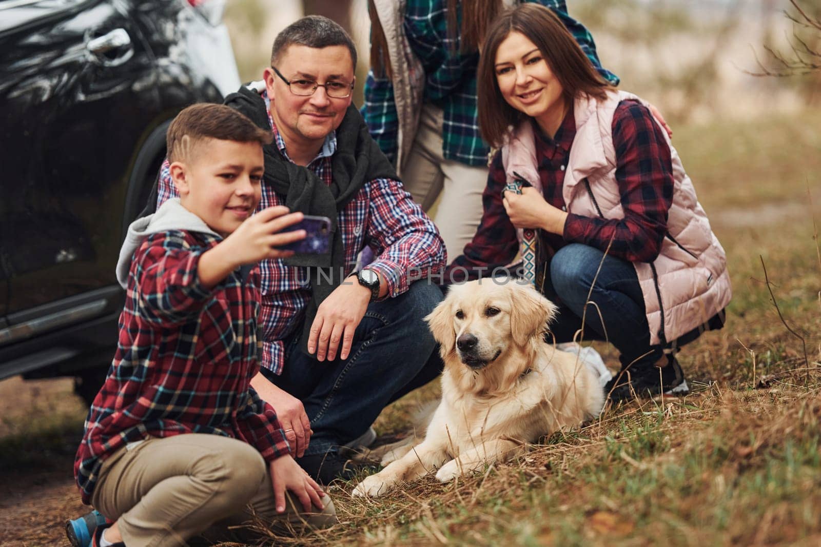 Happy family sitting and having fun with their dog near modern car outdoors in forest.