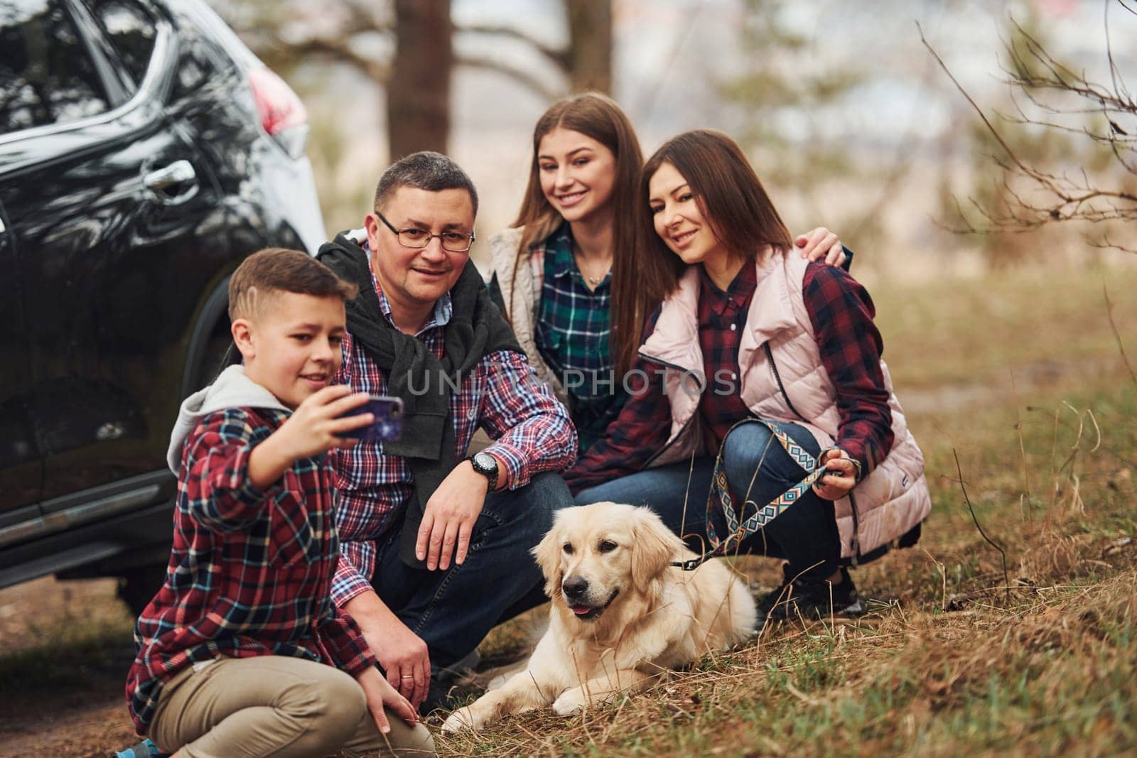 Happy family sitting and having fun with their dog near modern car outdoors in forest by Standret