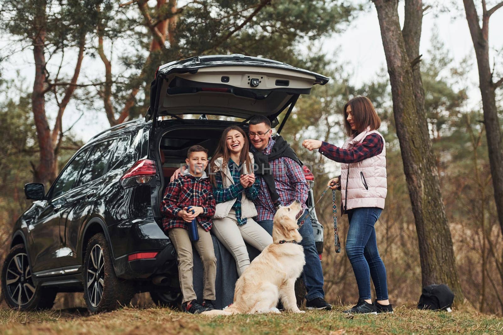 Happy family have fun with their dog near modern car outdoors in forest.