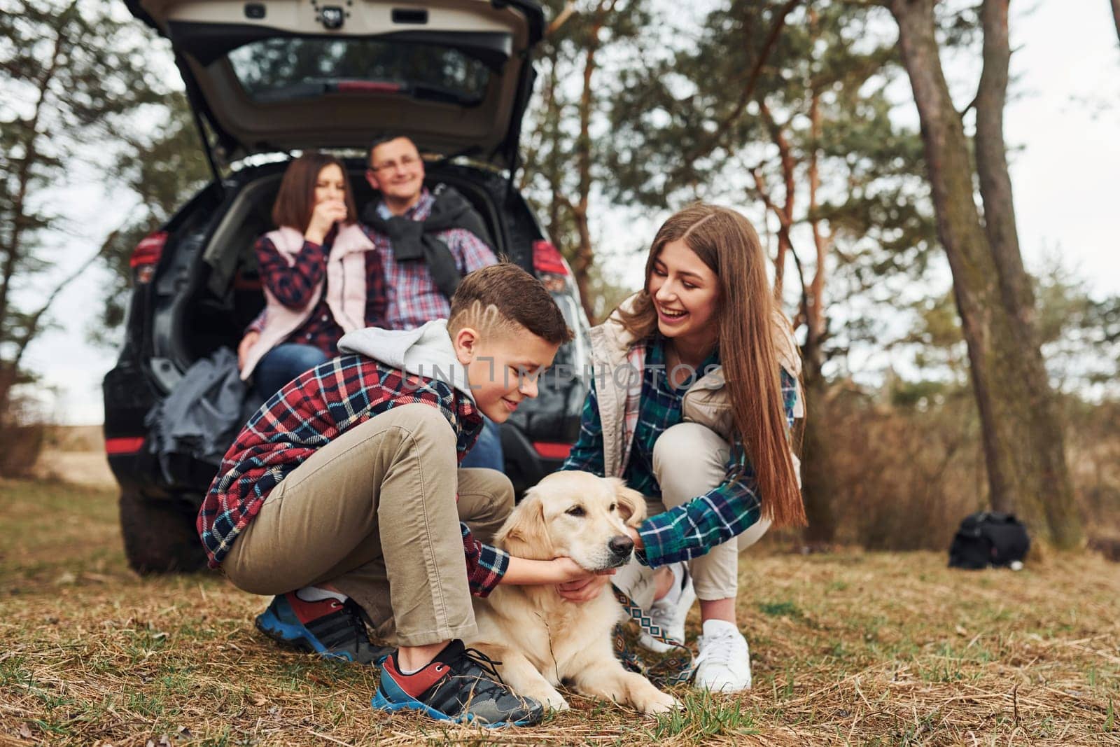 Happy family sitting and having fun with their dog near modern car outdoors in forest.