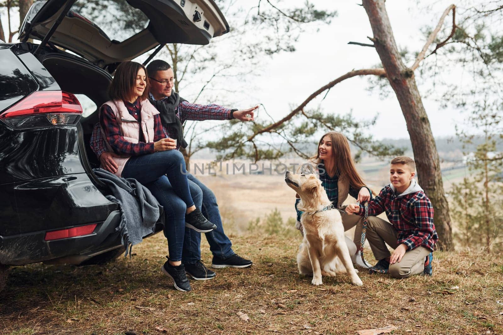 Happy family sitting and having fun with their dog near modern car outdoors in forest.