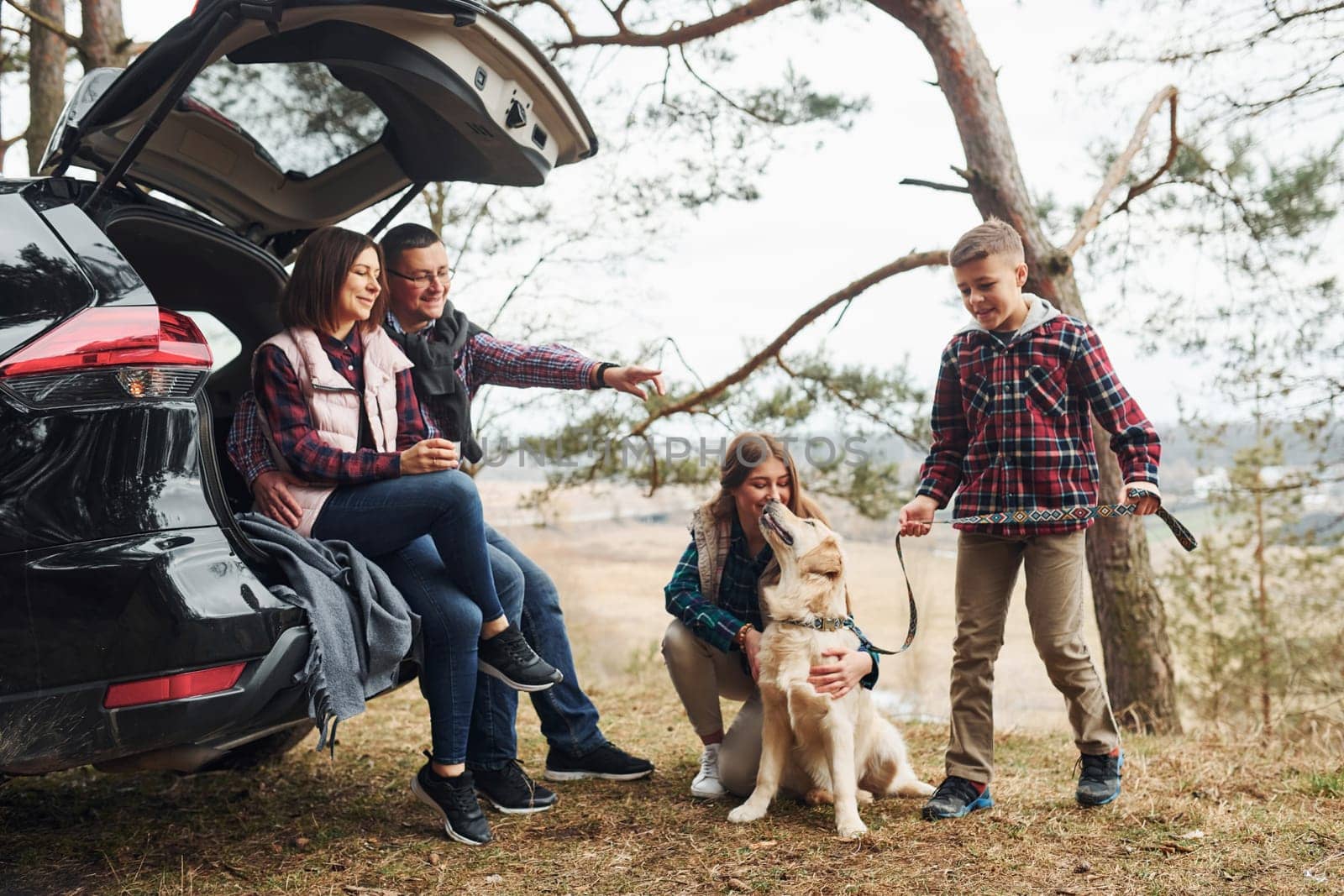 Happy family sitting and having fun with their dog near modern car outdoors in forest.