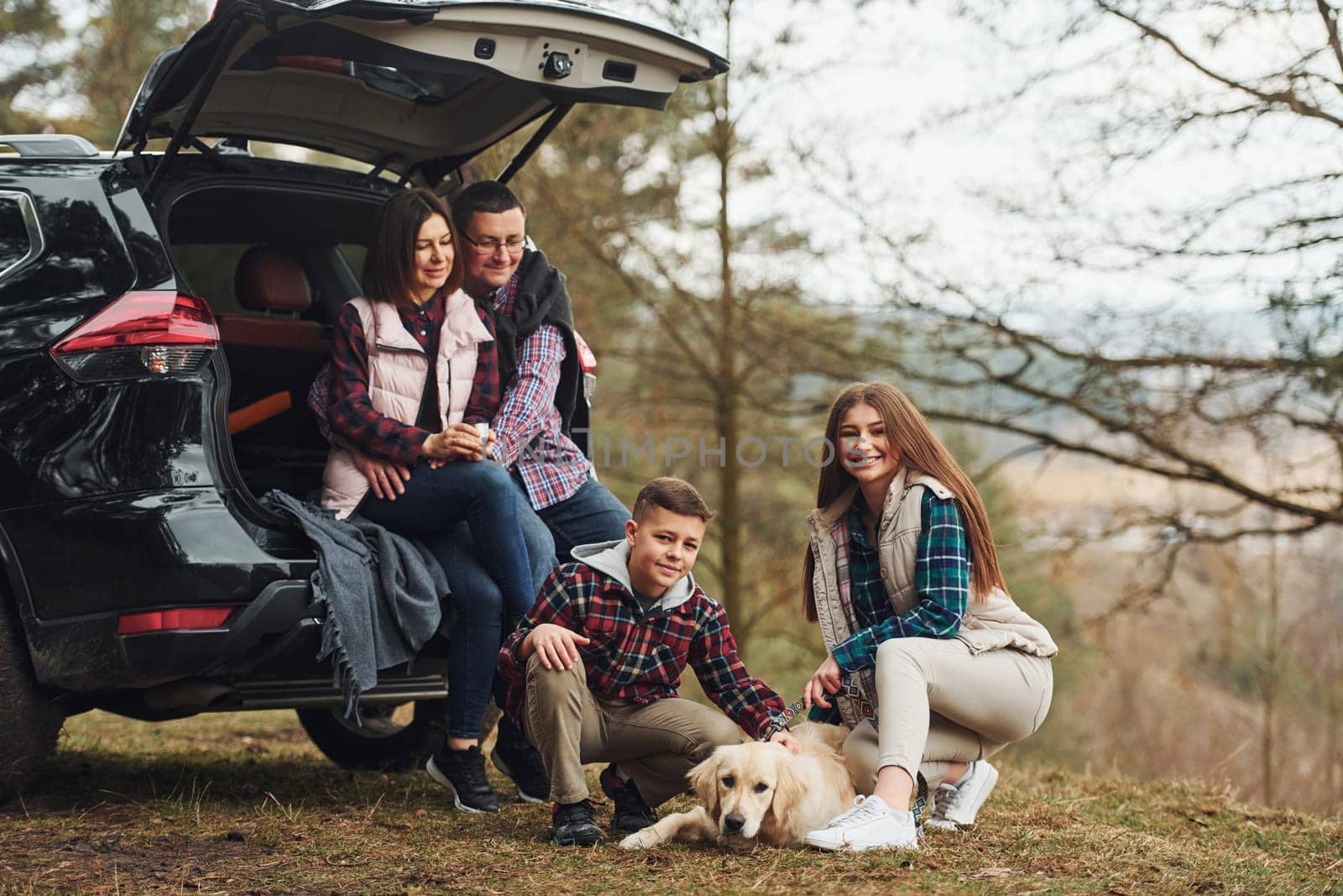 Happy family sitting and having fun with their dog near modern car outdoors in forest.