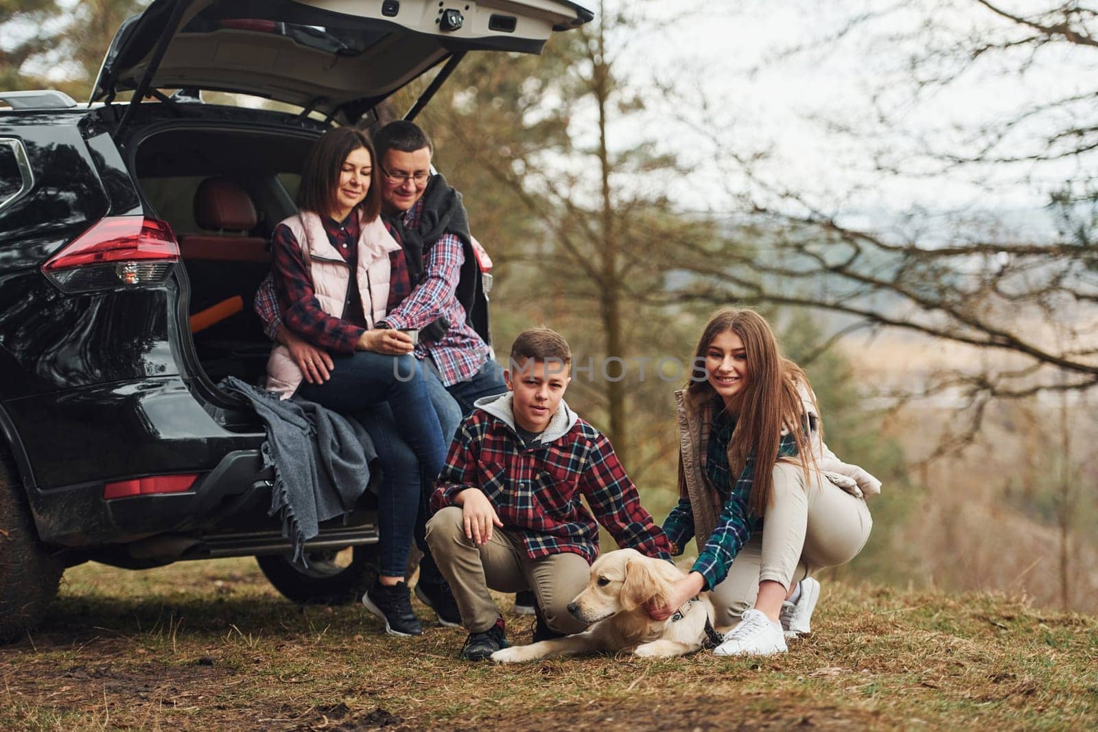 Happy family sitting and having fun with their dog near modern car outdoors in forest.