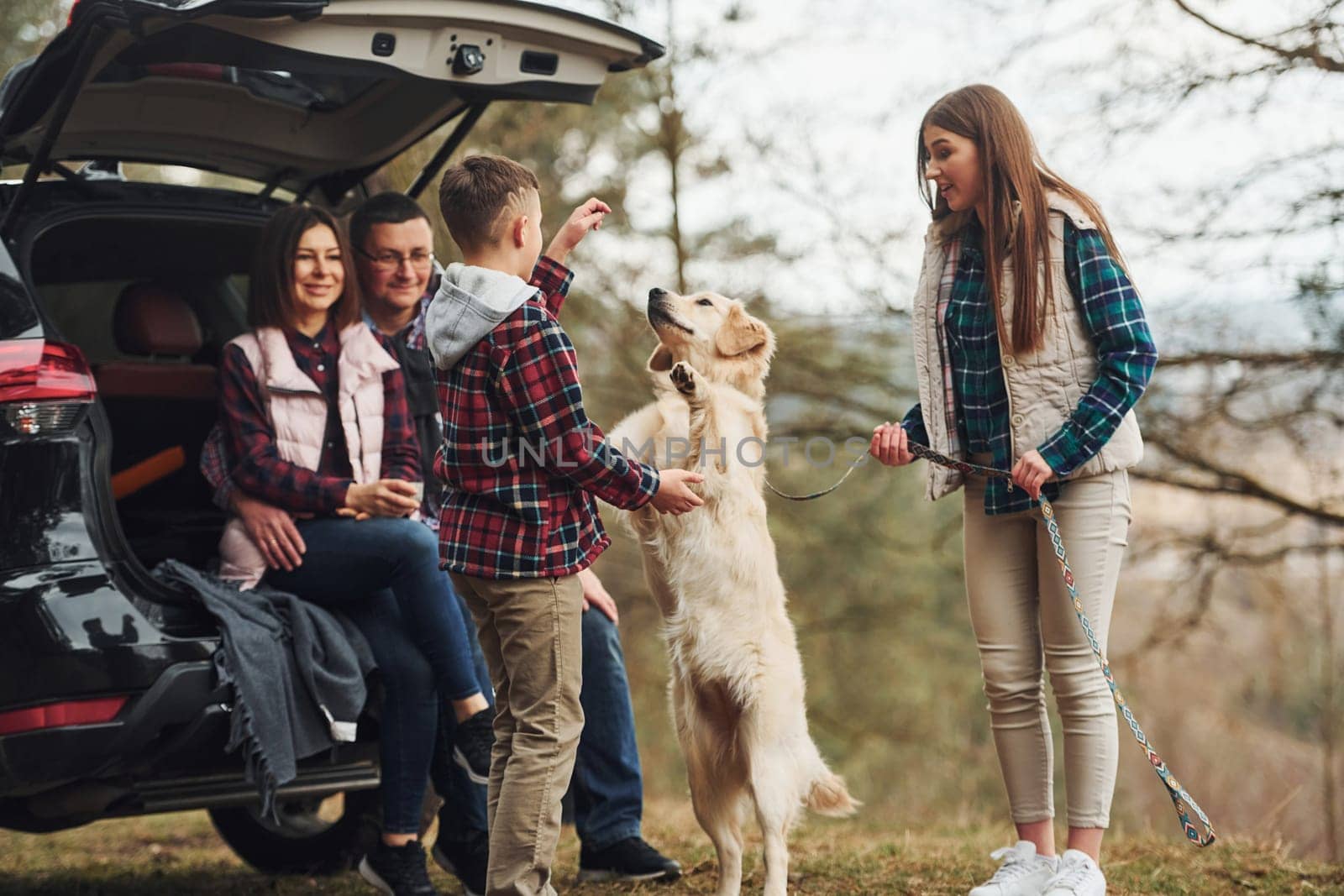 Happy family sitting and having fun with their dog near modern car outdoors in forest.