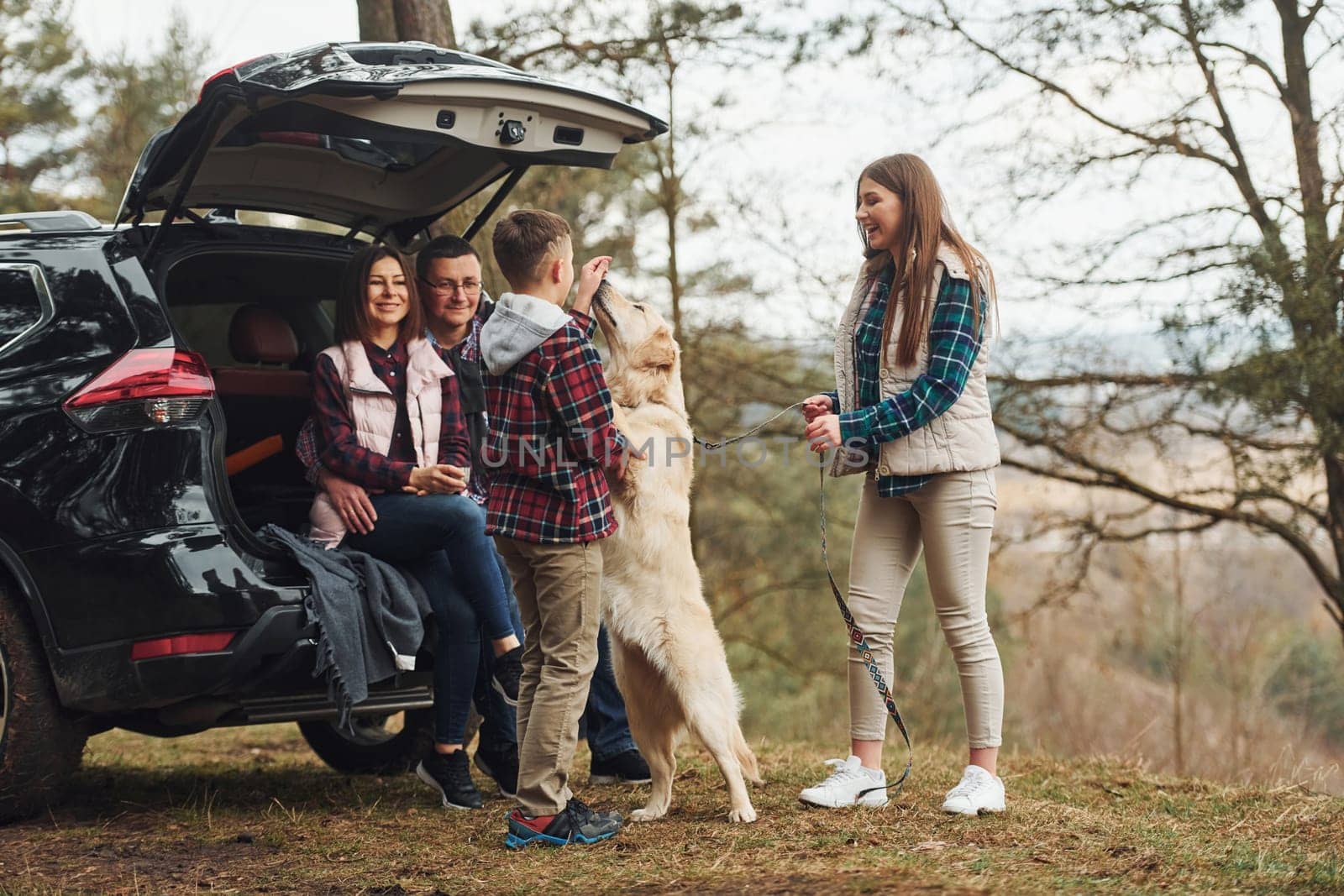 Happy family have fun with their dog near modern car outdoors in forest.