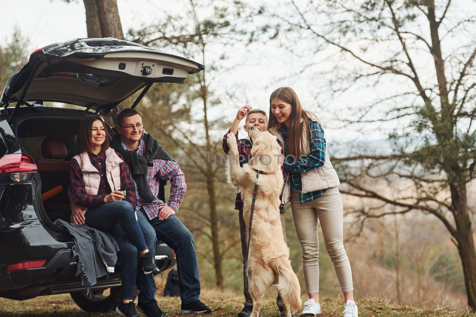 Happy family have fun with their dog near modern car outdoors in forest.