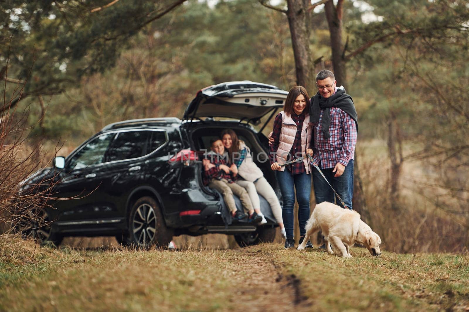 Happy family have fun with their dog near modern car outdoors in forest.