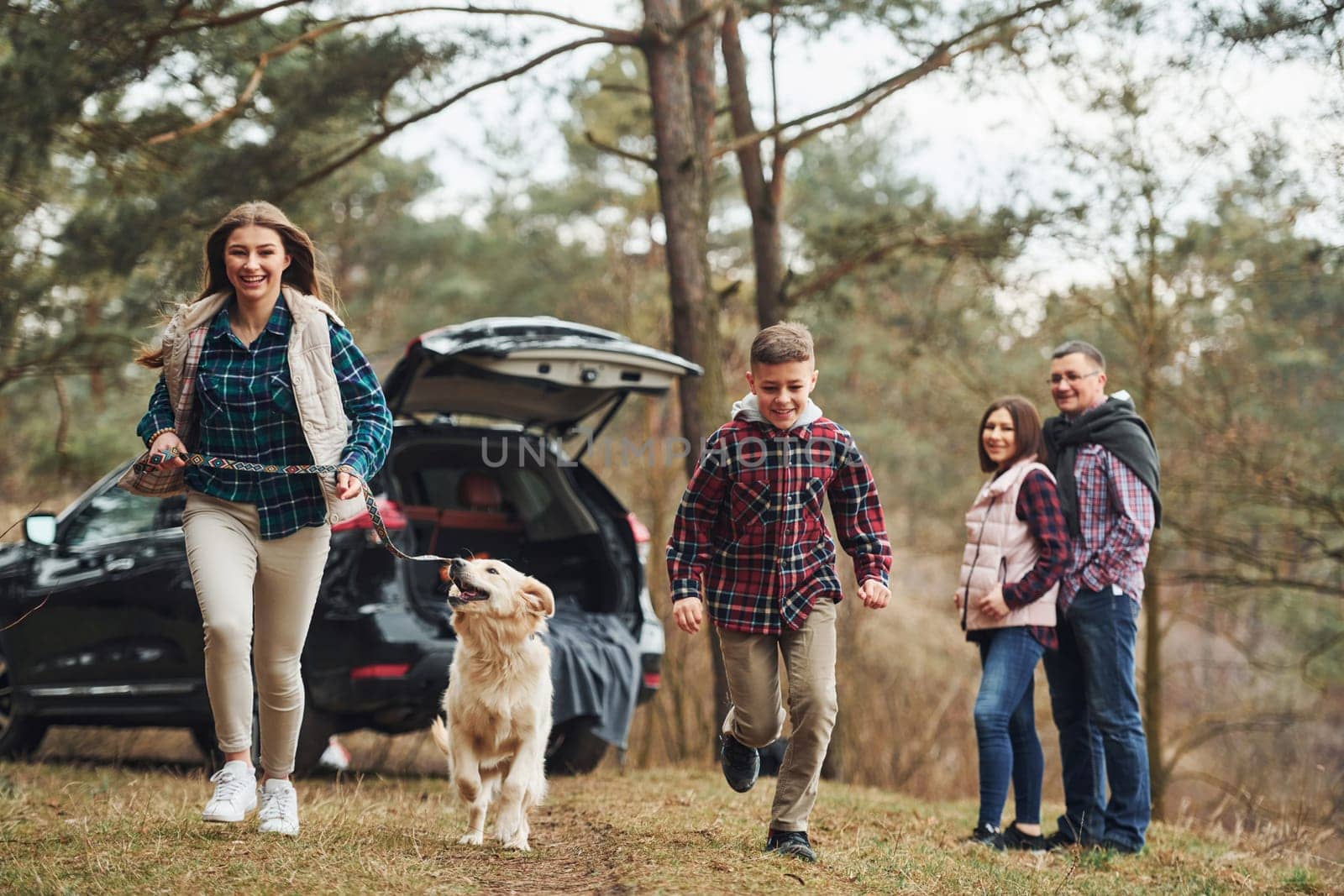 Sister and brother runs forward. Happy family have fun with their dog near modern car outdoors in forest by Standret
