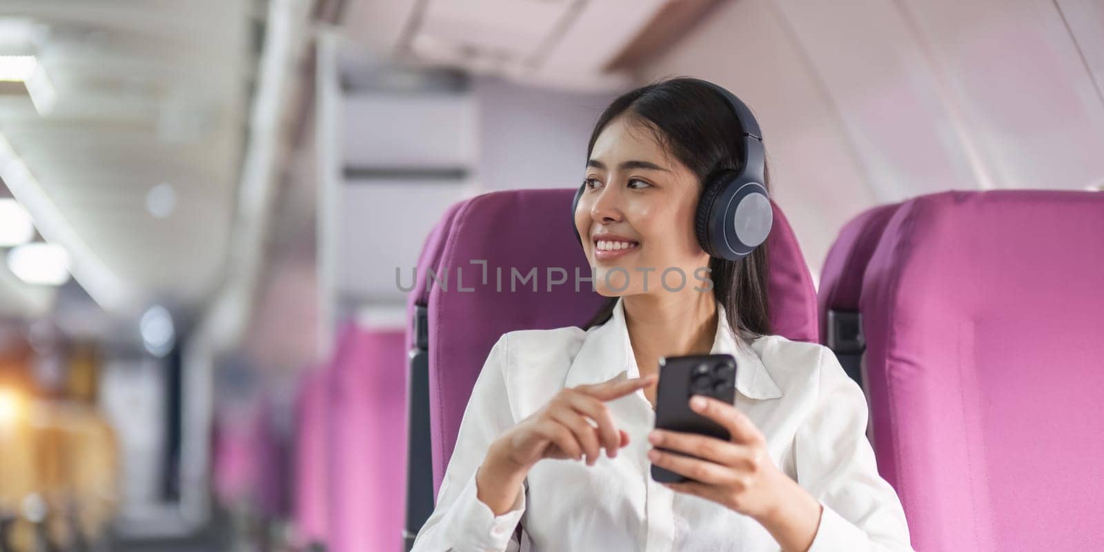 Cheerful female passenger in headphones for noise cancellation watching online movie during intercontinental flight in cabin of aircraft, happy young woman using wifi connection on board by nateemee