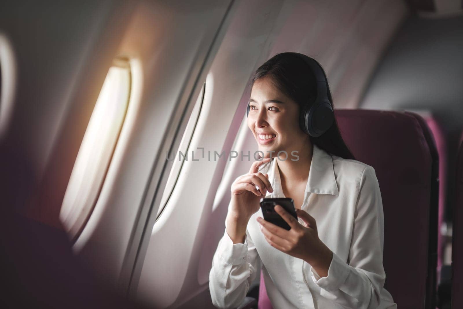 Cheerful female passenger in headphones for noise cancellation watching online movie during intercontinental flight in cabin of aircraft, happy young woman using wifi connection on board by nateemee
