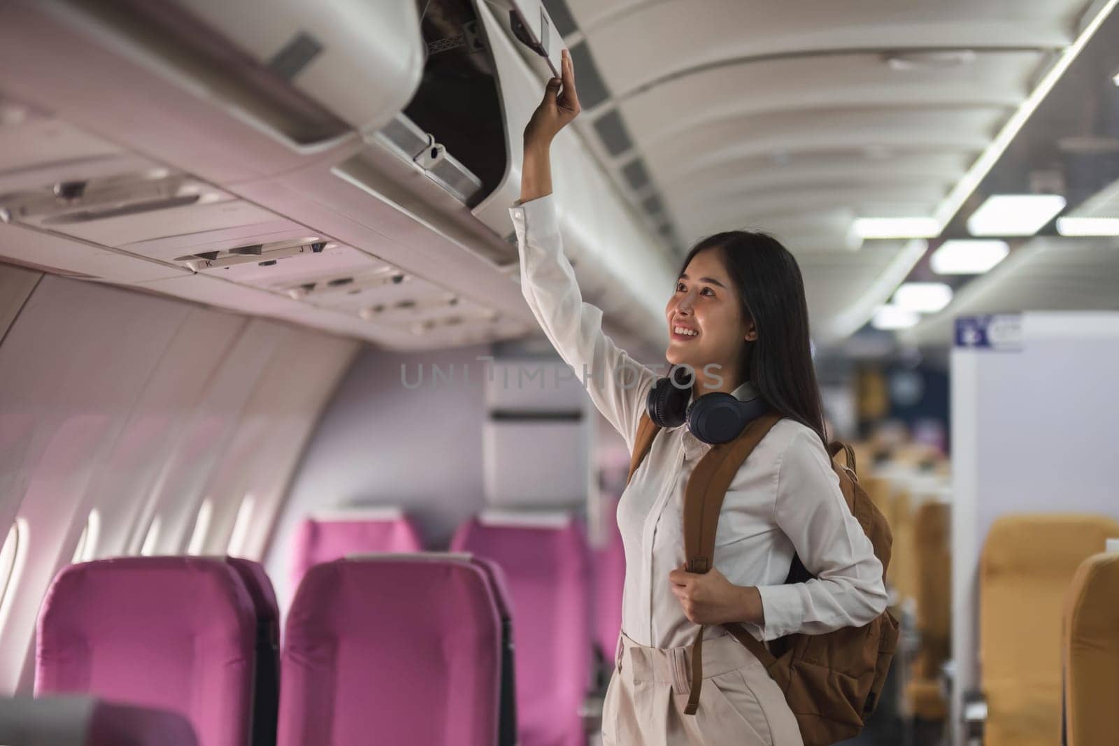 Young asian woman travel by airplane. Passenger putting hand baggage in lockers above seats of plane.