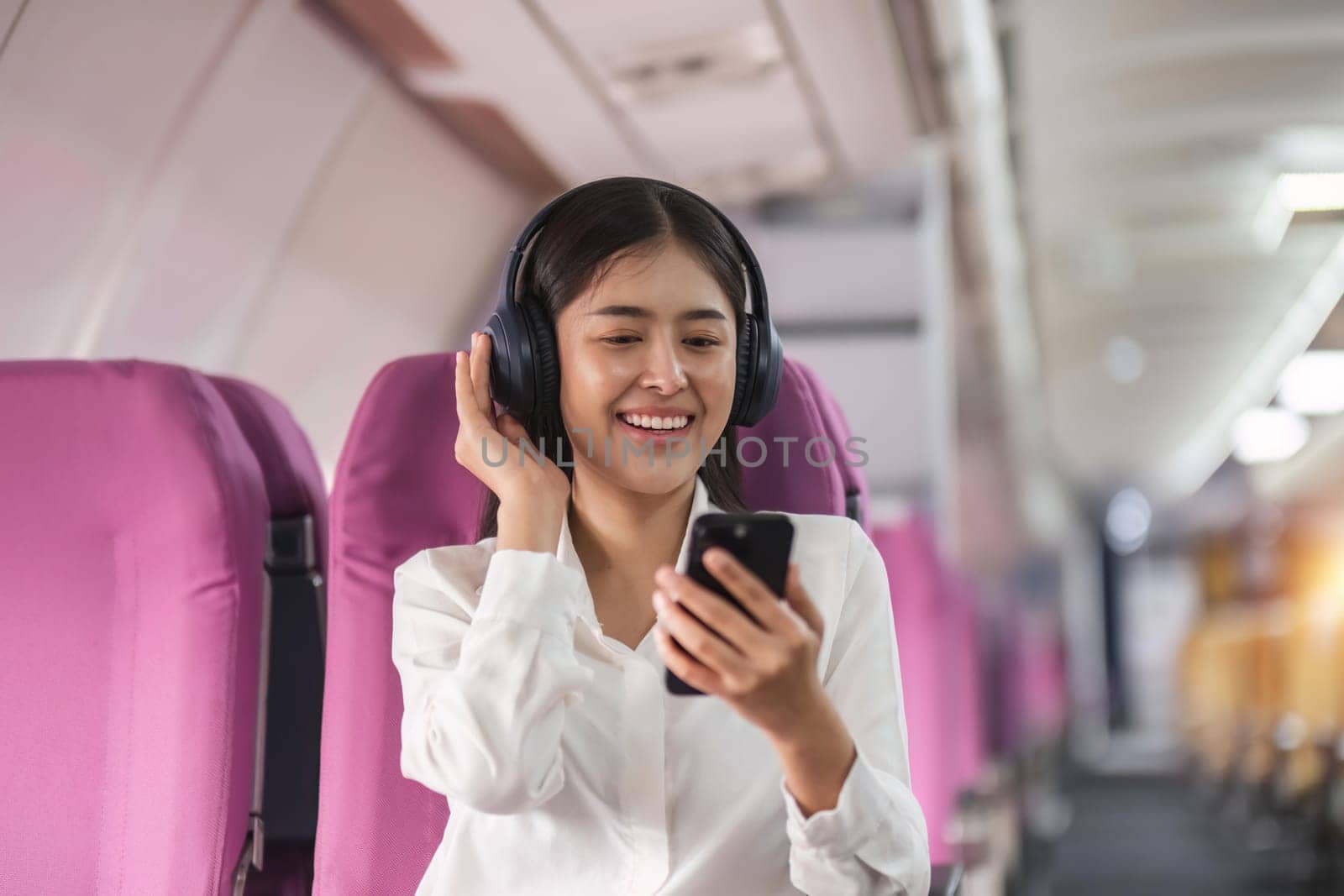 Cheerful female passenger in headphones for noise cancellation watching online movie during intercontinental flight in cabin of aircraft, happy young woman using wifi connection on board.