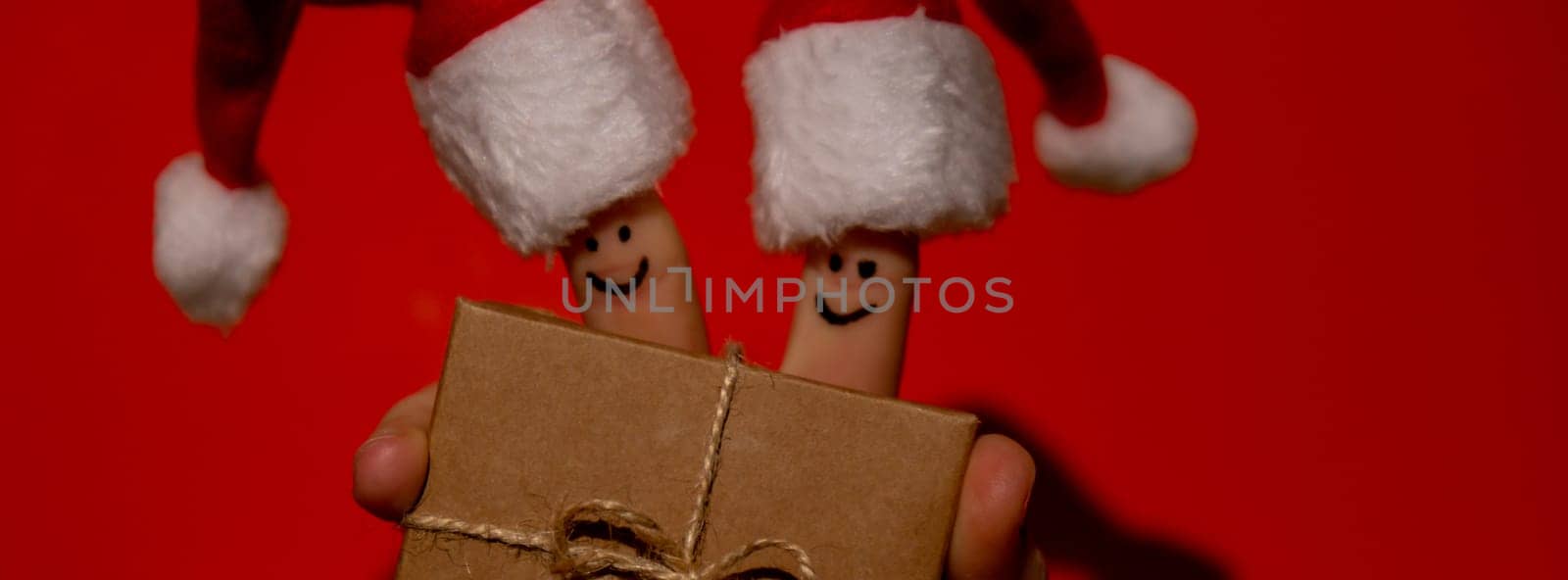 Fingers dressed in Santa-Claus red-white hats. Two happy finger smileys faces holding paper gift box present against red background. Happy family celebrating concept for Christmas or New Years day. couple by anna_stasiia