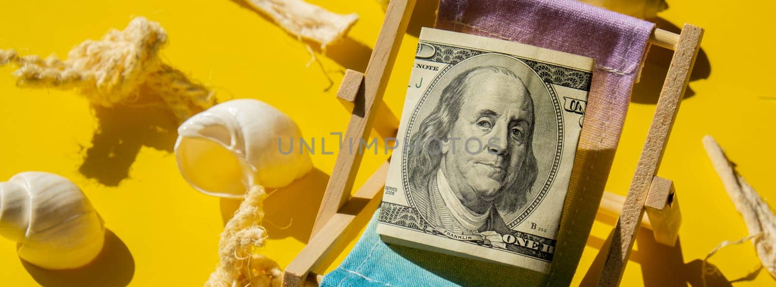 Rolled Up Hundred Dollar Banknote On Miniature Deck beach Chair. Concept of costs in travel holidays. Save for vacation. Copy space. The concept of how much it costs to rest Banner