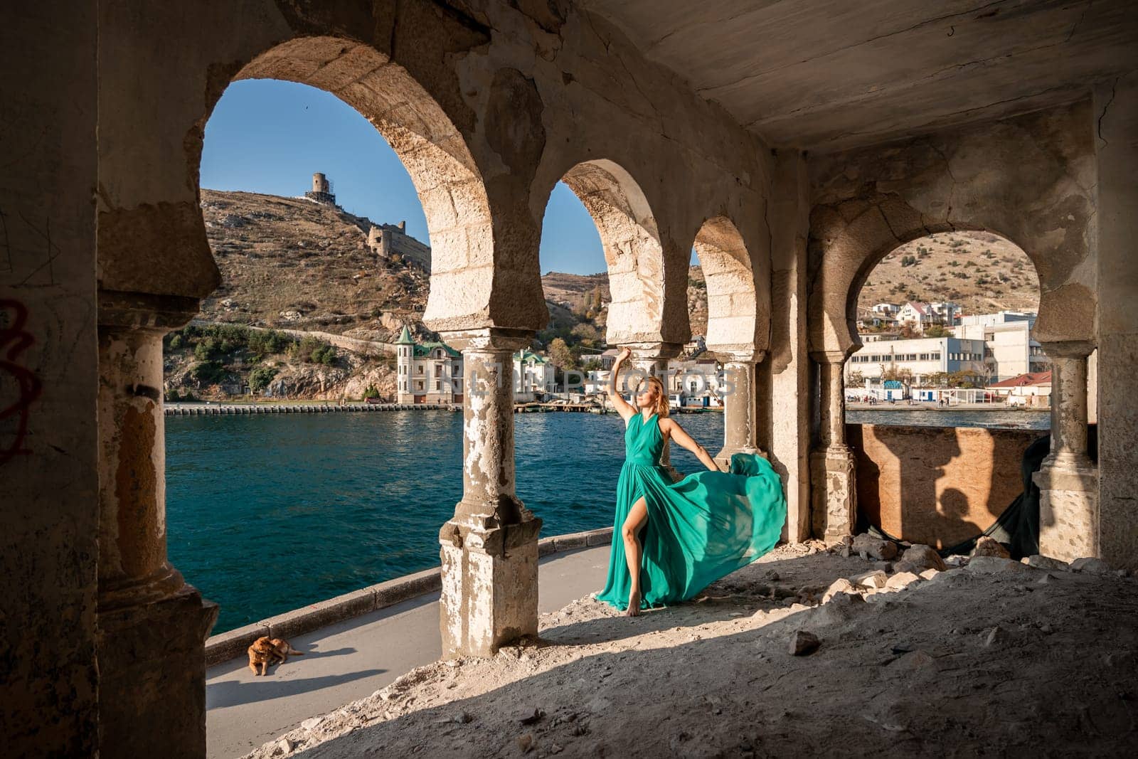 Happy blonde in a long mint dress posing against the backdrop of the sea in an old building with columns. Girl in nature against the blue sky