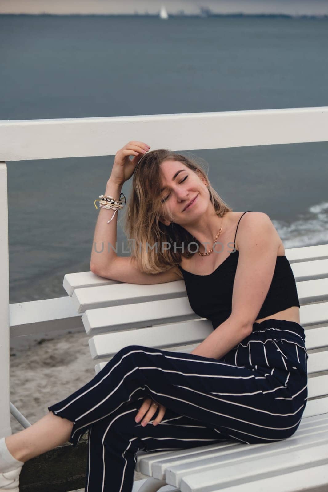 Young woman sitting on bench on wooden pier blurred beachside background. Attractive female enjoying the sea shore travel and active lifestyle concept. Springtime. Wellness wellbeing mental health inner peace Slow life digital detox