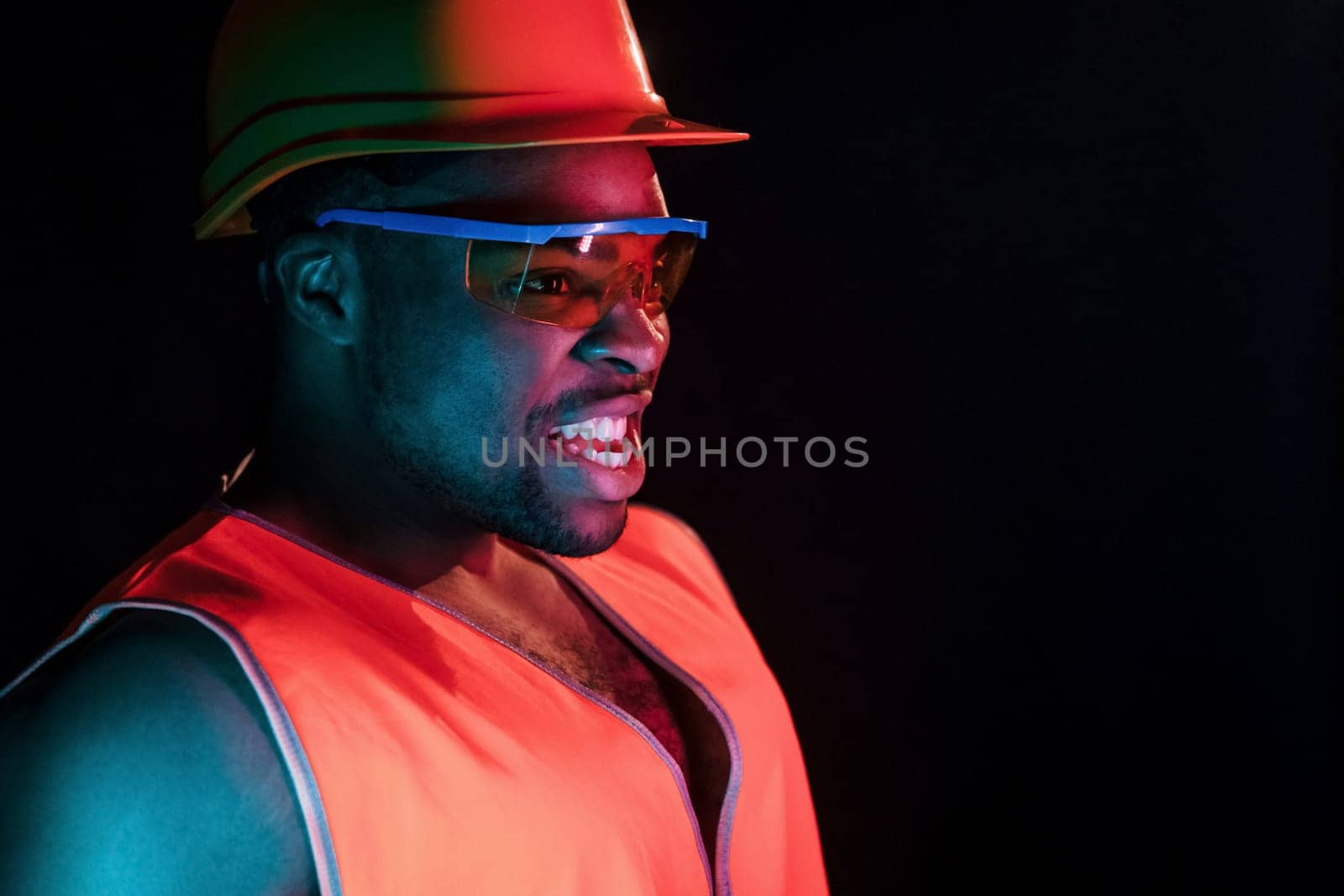 Construction worker in uniform and hard hat. Futuristic neon lighting. Young african american man in the studio by Standret