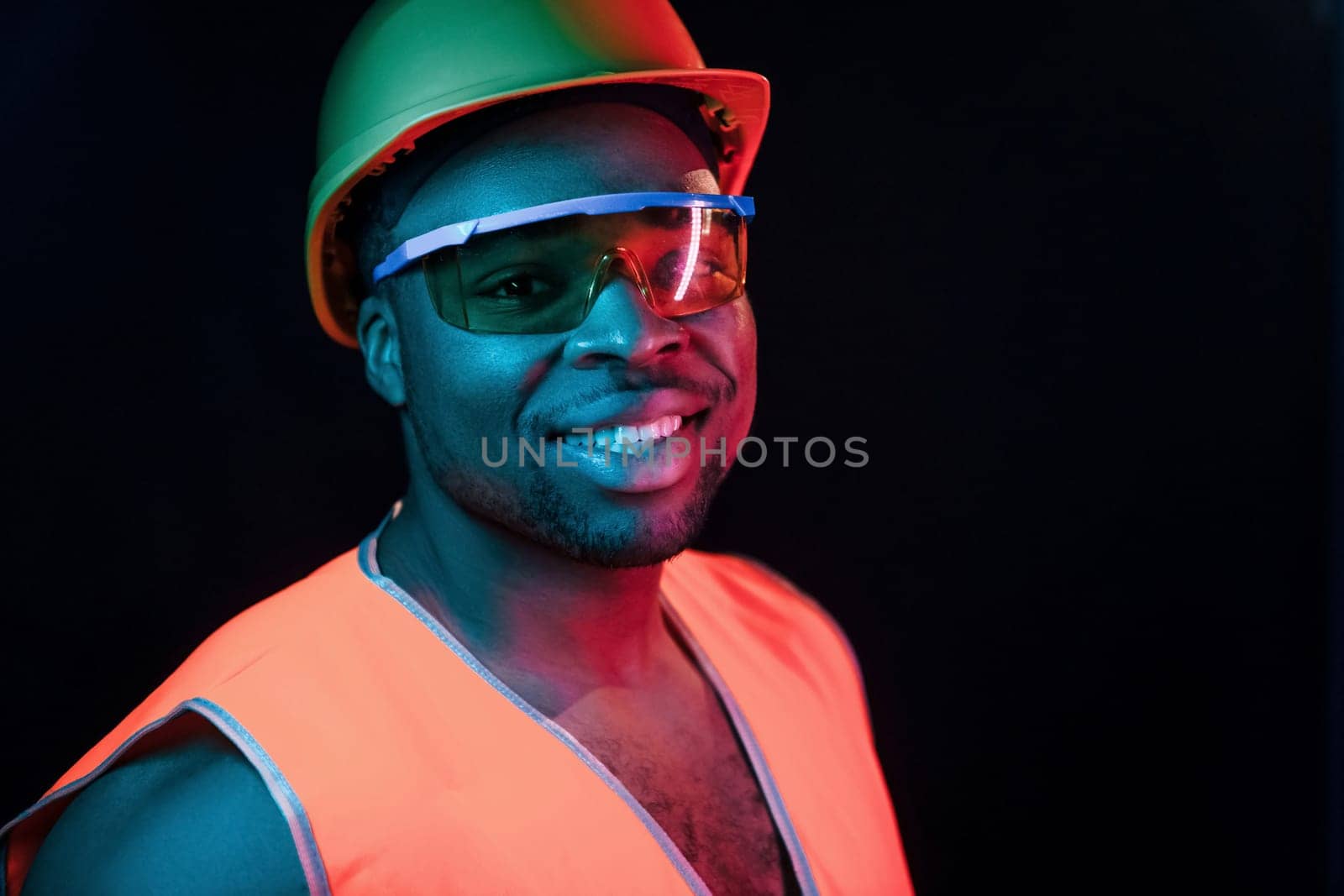 Construction worker in uniform and hard hat. Futuristic neon lighting. Young african american man in the studio by Standret