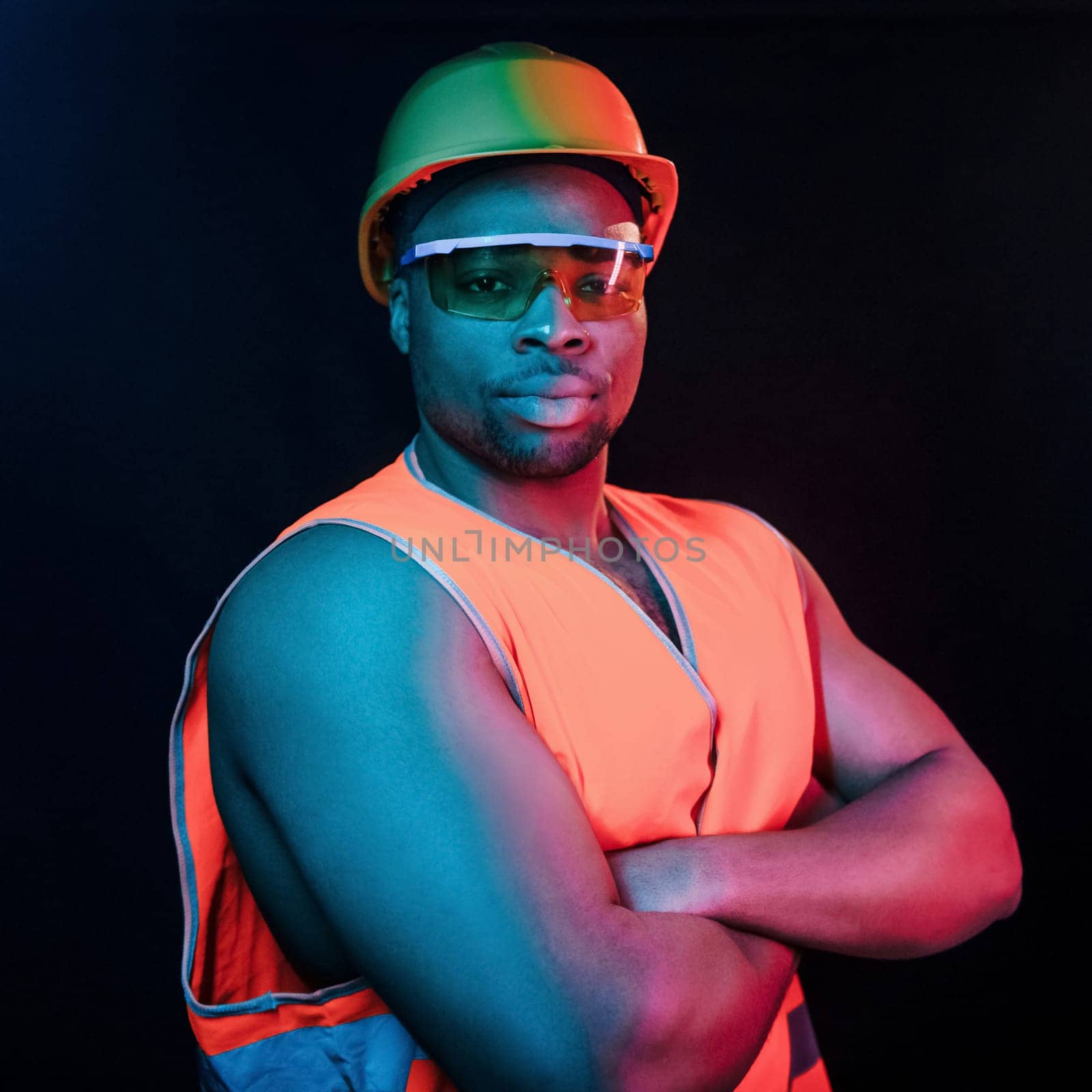 Construction worker in uniform and hard hat. Futuristic neon lighting. Young african american man in the studio.