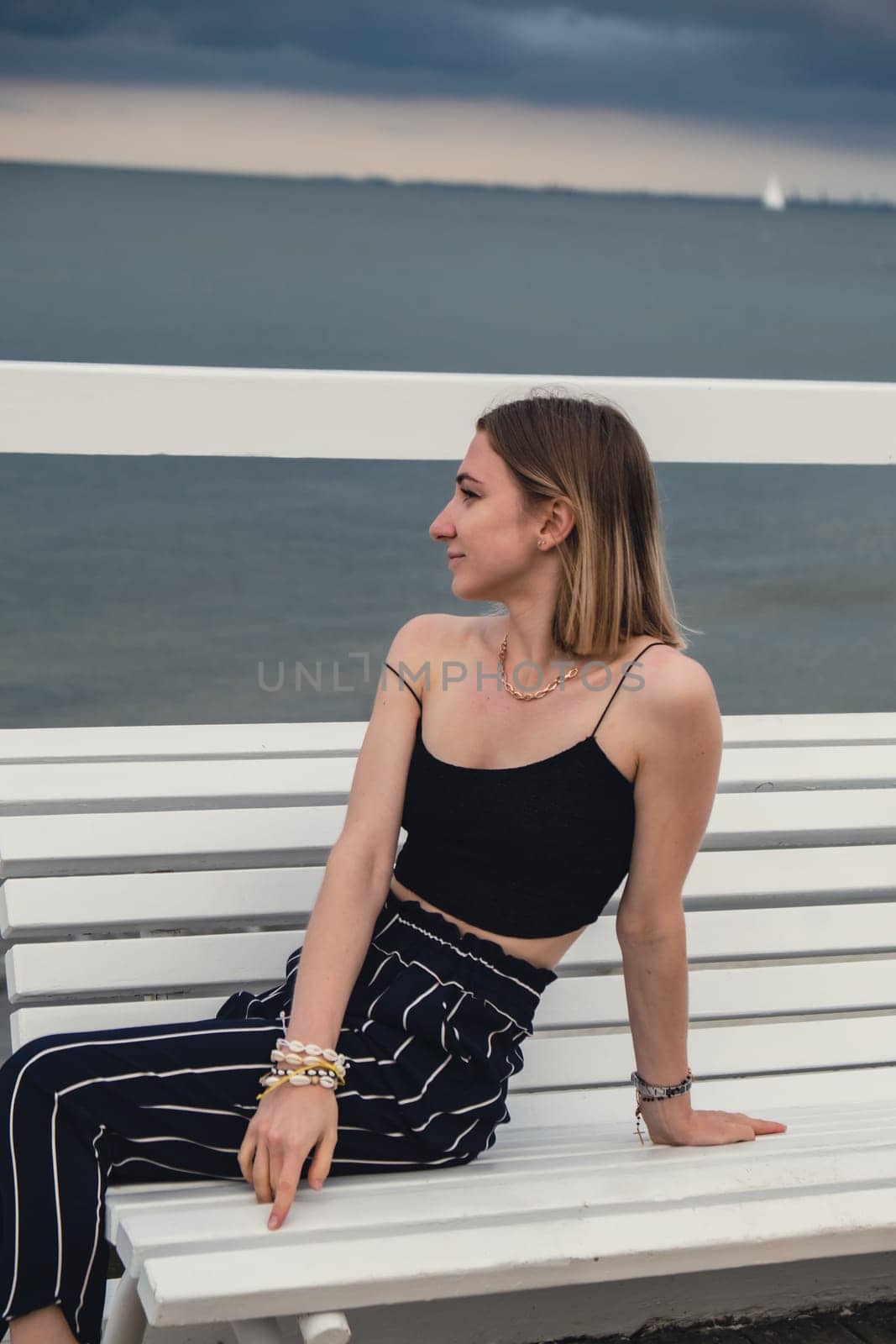 Young woman sitting on bench on wooden pier blurred beachside background. Attractive female enjoying the sea shore travel and active lifestyle concept. Springtime. Wellness wellbeing mental health inner peace Slow life digital detox