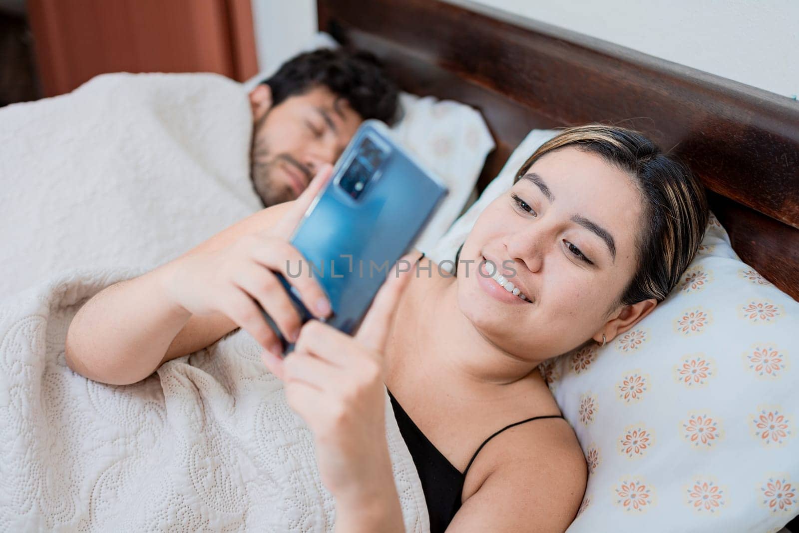 Unfaithful wife with phone while her husband sleeps. Unfaithful woman with phone while the man sleeps. Unfaithful girlfriend with phone while boyfriend sleeps by isaiphoto