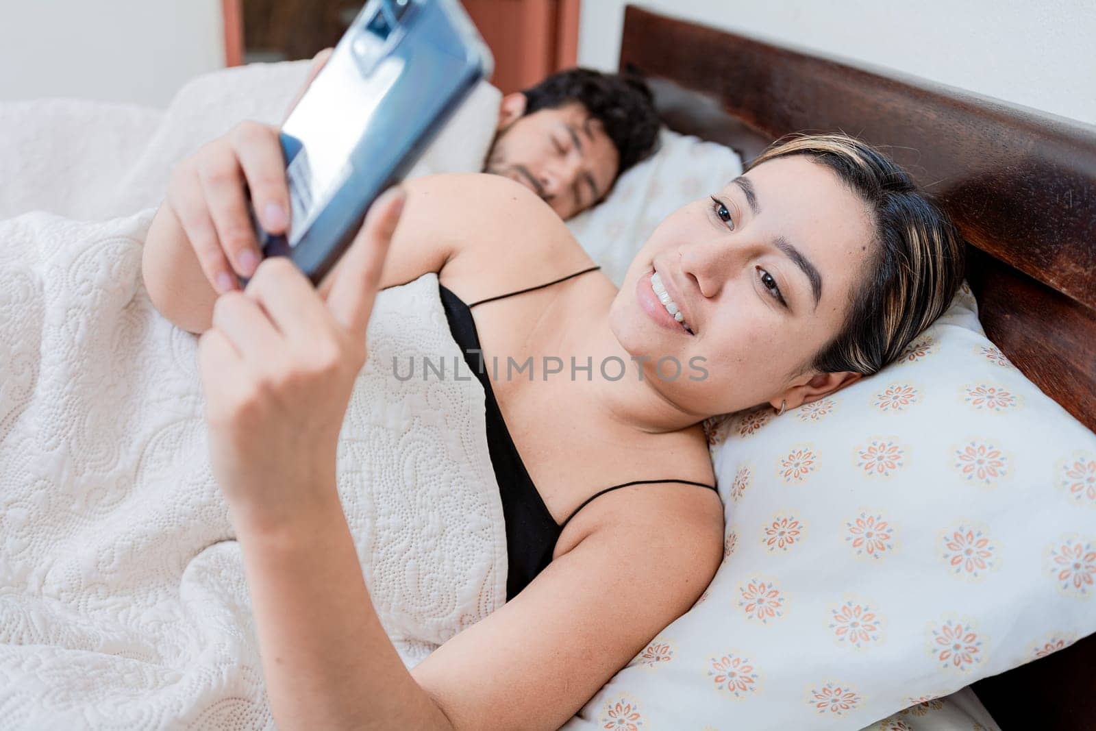 Unfaithful woman with phone while the man sleeps. Unfaithful girlfriend with phone while boyfriend sleeps, Unfaithful wife with phone while her husband sleeps