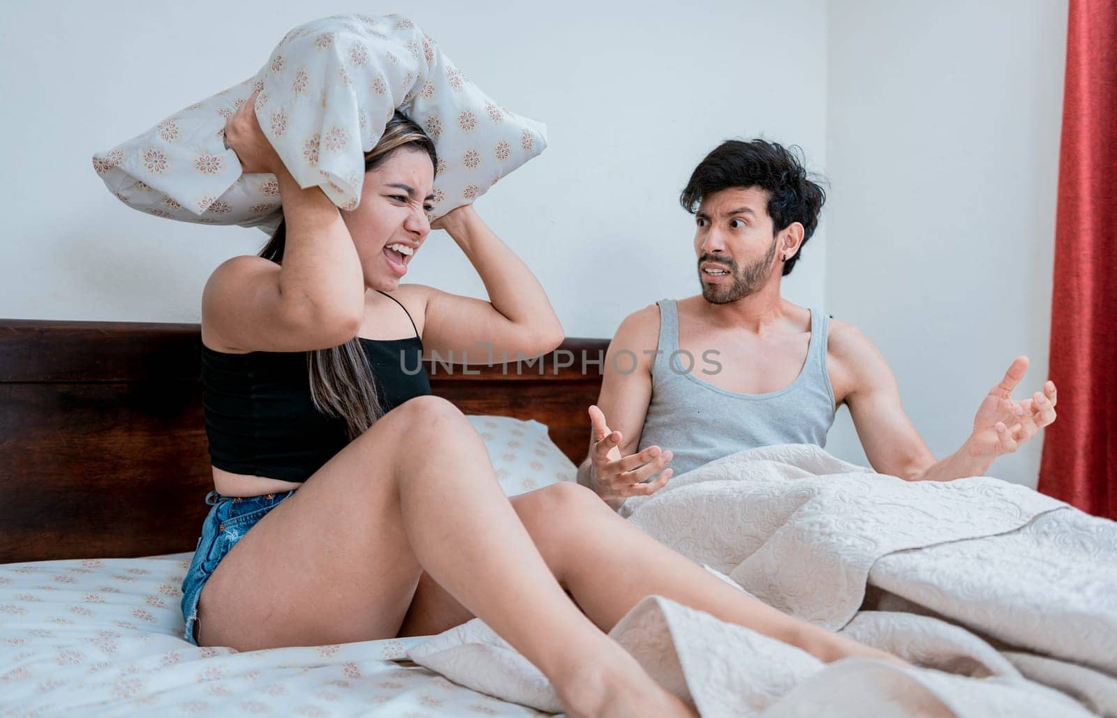 Young couple arguing in bed with the woman screaming and the man covering his ears with a pillow. Upset woman yelling at husband in bedroom bed, Couple arguing in bed room by isaiphoto