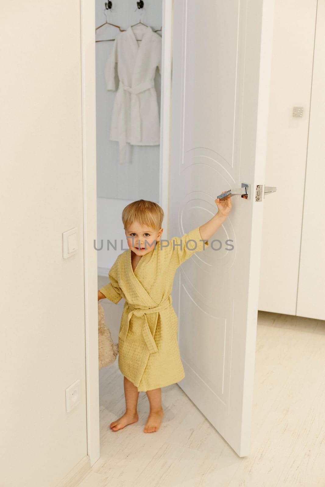 A boy in a yellow bathrobe stands at the entrance to the bathroom, smiling.