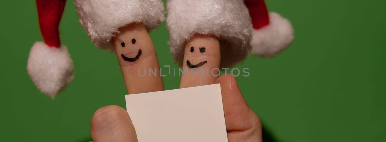 Fingers dressed in Santa-Claus red-white hats. Two happy finger smileys faces holding empty paper note for your text copy space against green background. Happy family celebrating concept for Christmas or New Years day. couple by anna_stasiia